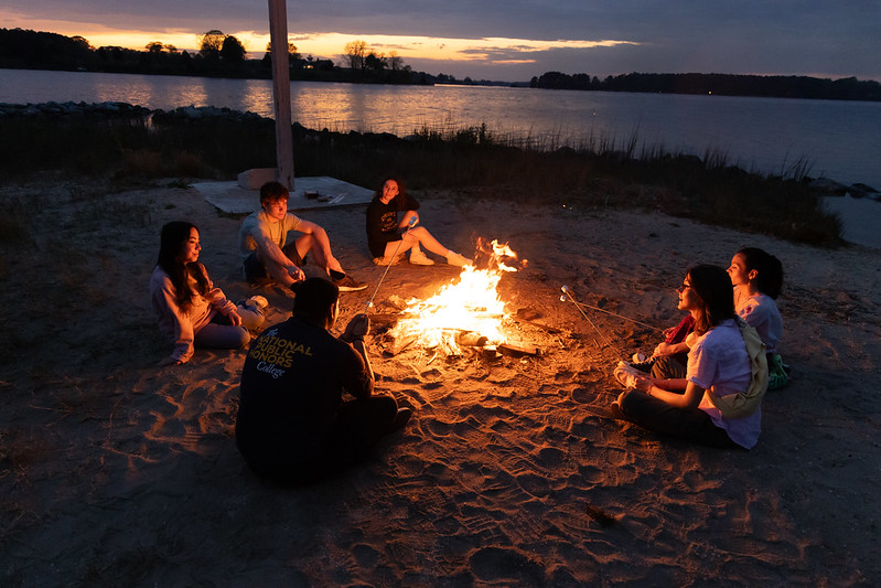 A calm moment with a beautiful sunset in the background with college students surrounding a camp fire roasting marshmallows.