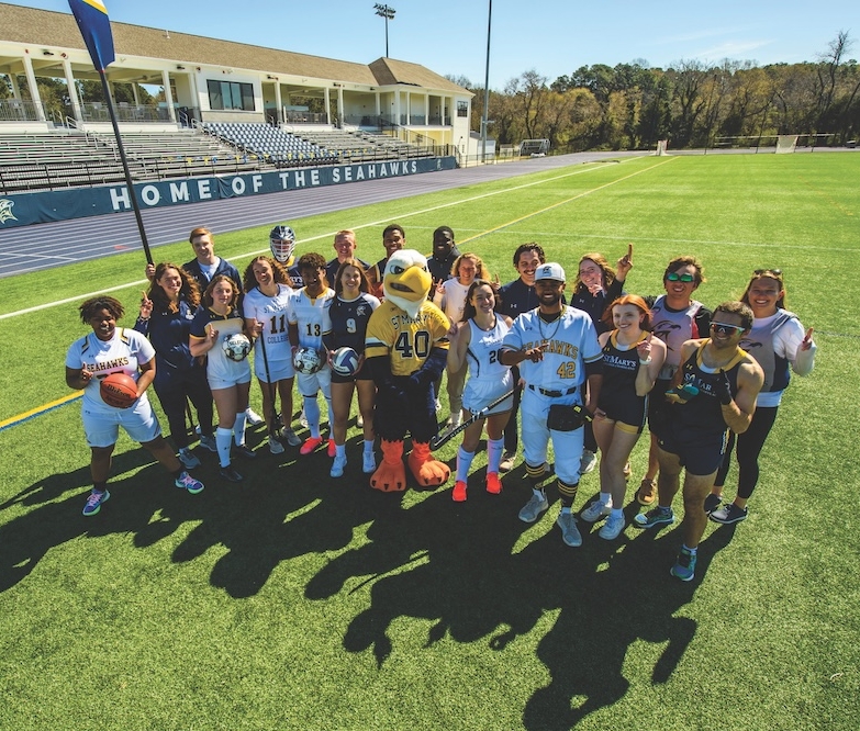 A diverse group of athletes in sports uniforms poses with a bird mascot on a soccer field. They stand in front of stadium seating labeled "Home of the Seahawks" on a sunny day.